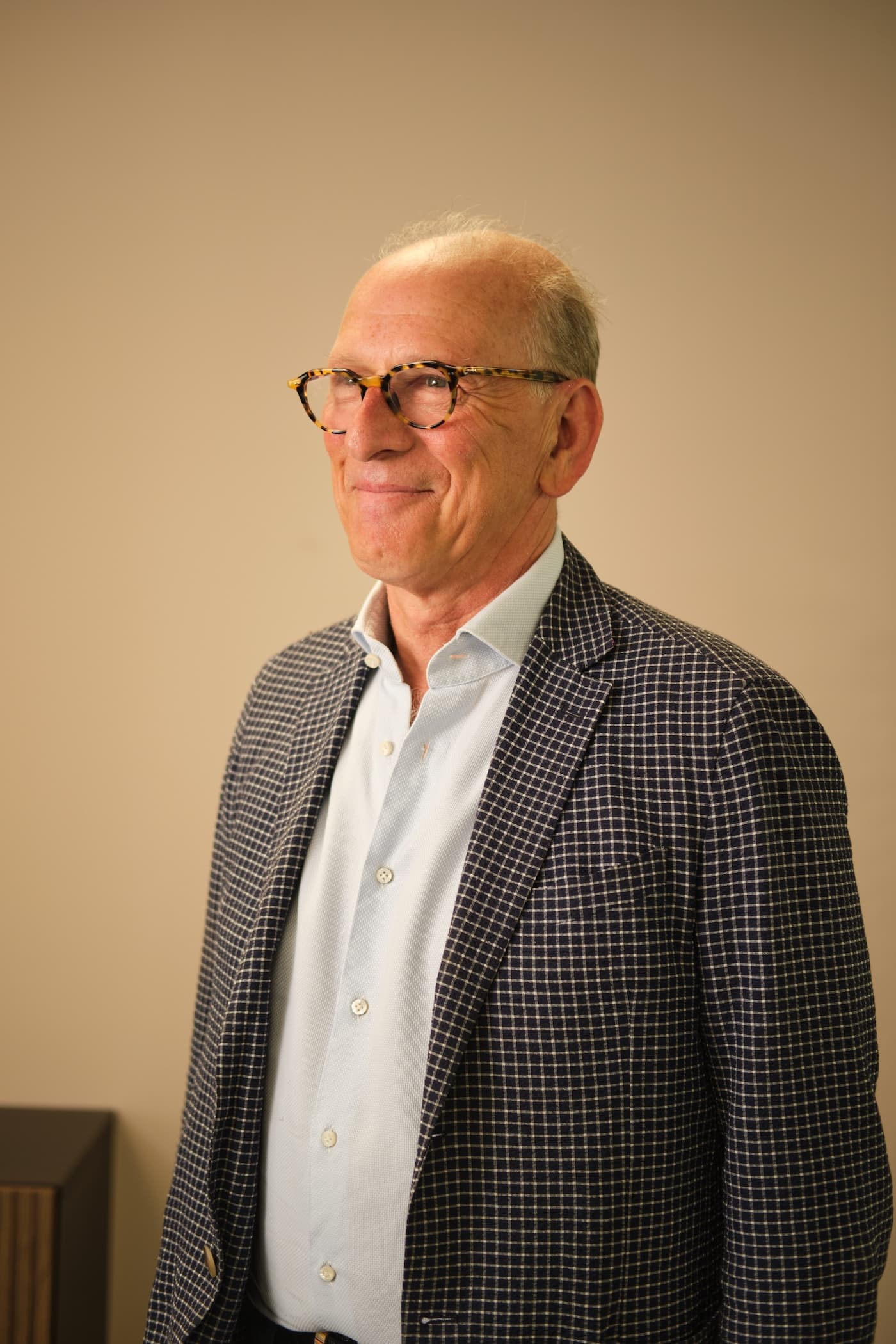 The image shows a person wearing a patterned blazer, a light-colored shirt, and glasses. The background is plain and the individual is looking slightly to the side with a smile.