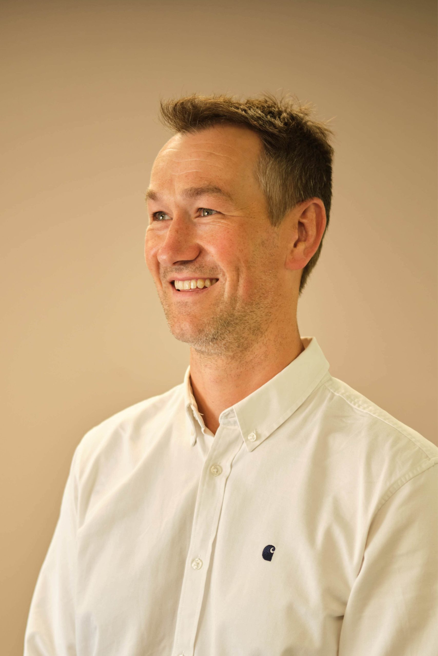 The image shows a man smiling, wearing a white button-up shirt against a neutral background.
