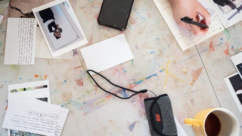 A hand writes a caption for a photo with a pen on a table where various printed photos and captions on cards lie, with a camera and a cup of tea.