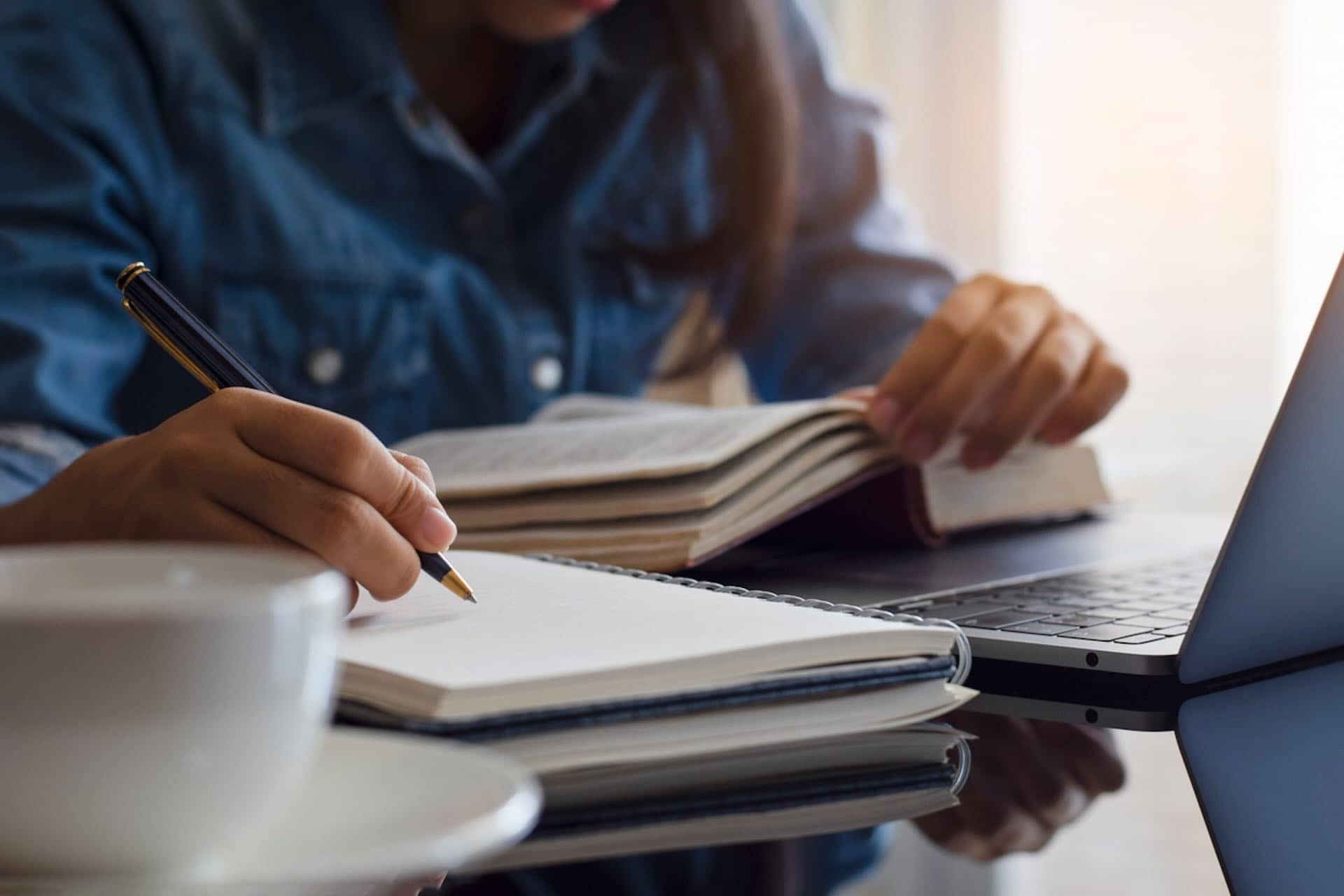 The image shows a person studying at a desk. They're writing in a notebook and have an open book. A laptop and a cup of coffee or tea are also present on the table. The setting suggests a focused study or work environment.