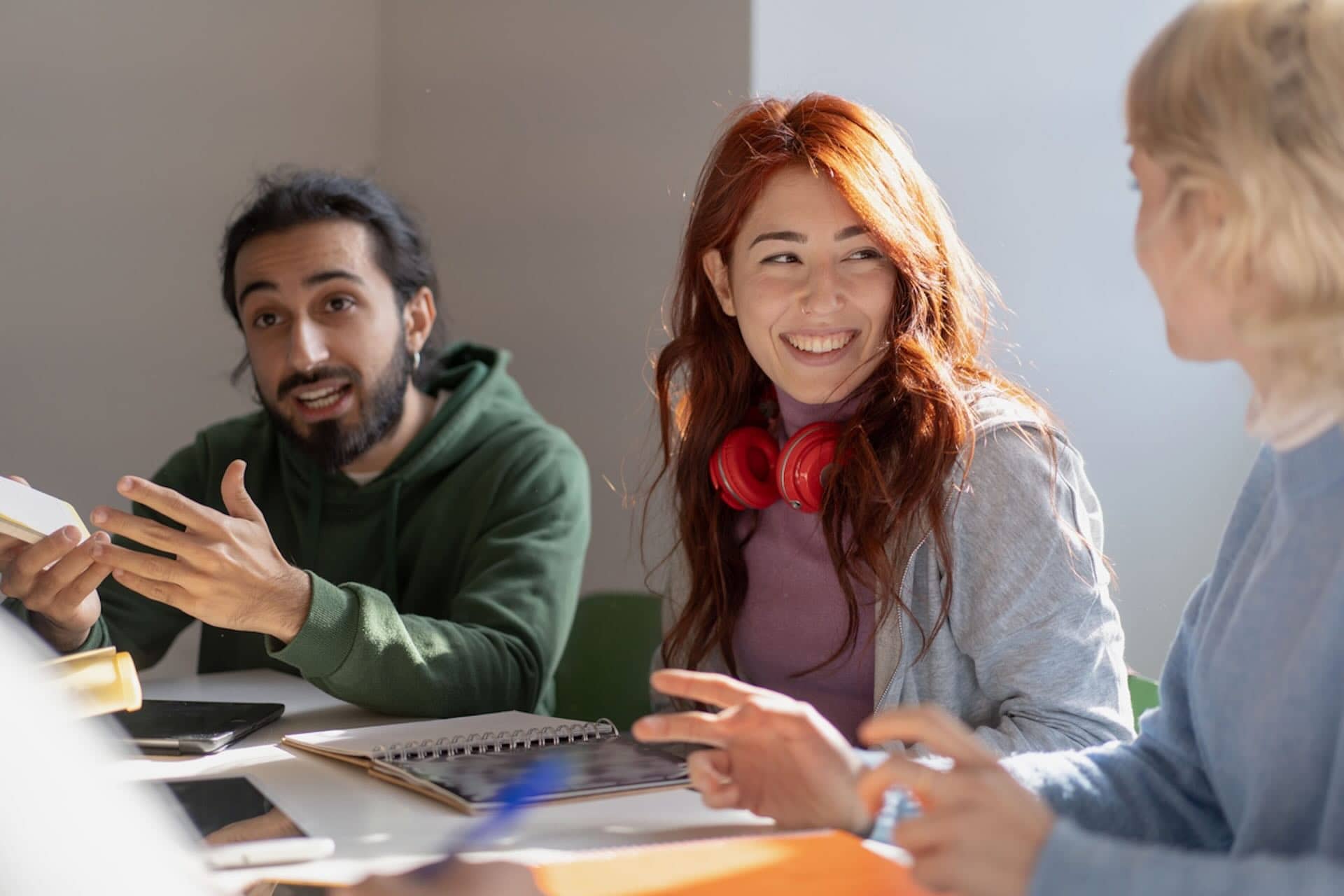 The image shows three people sitting at a table engaged in a conversation. One person on the left, wearing a green hoodie, seems to be speaking, while the person in the middle with red hair and red headphones is smiling. The person on the right