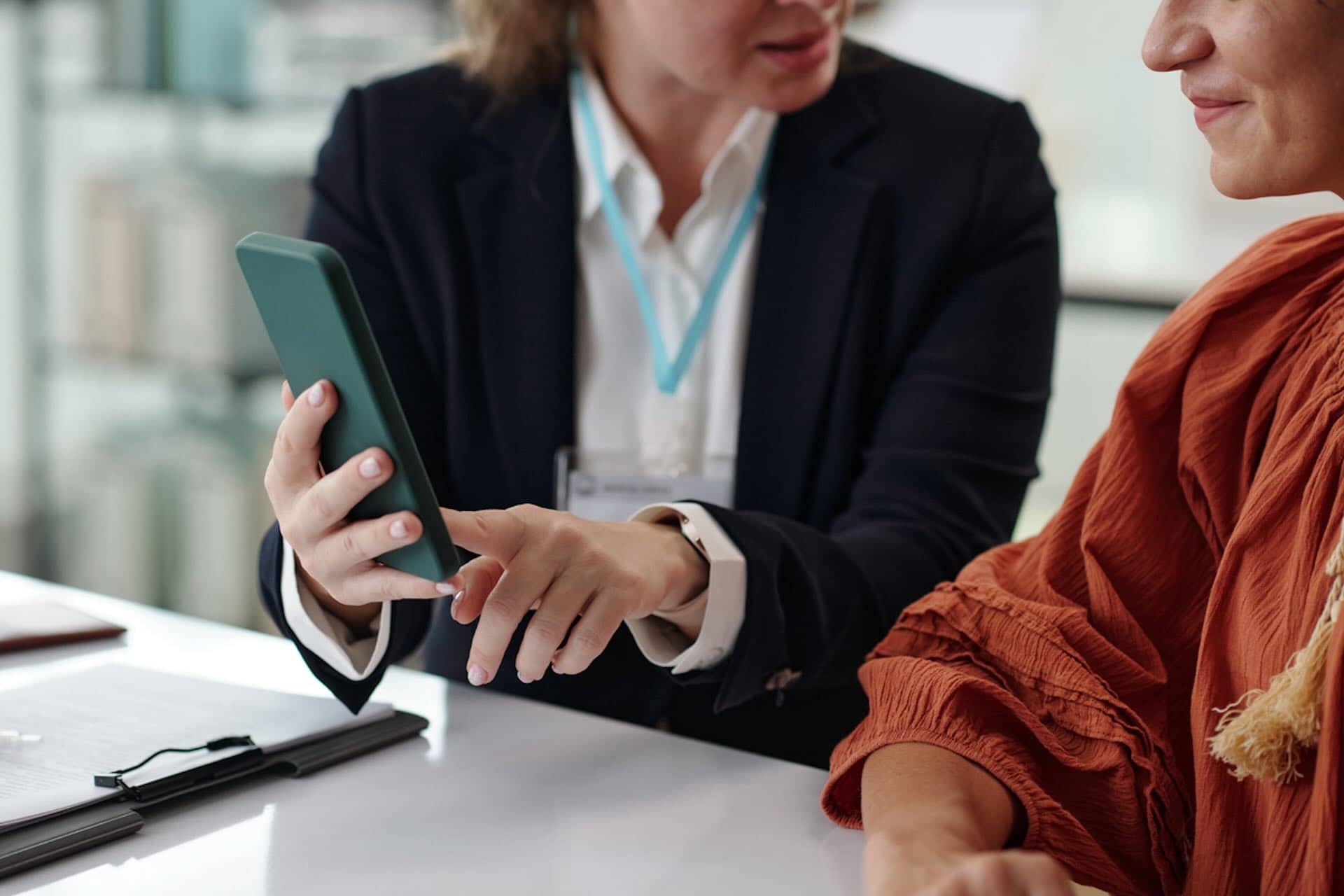 The image shows two people in a conversation. One person is holding a smartphone and pointing at it, appearing to show something to the other person. They are sitting at a table with some documents and a lanyard is visible on one of them.