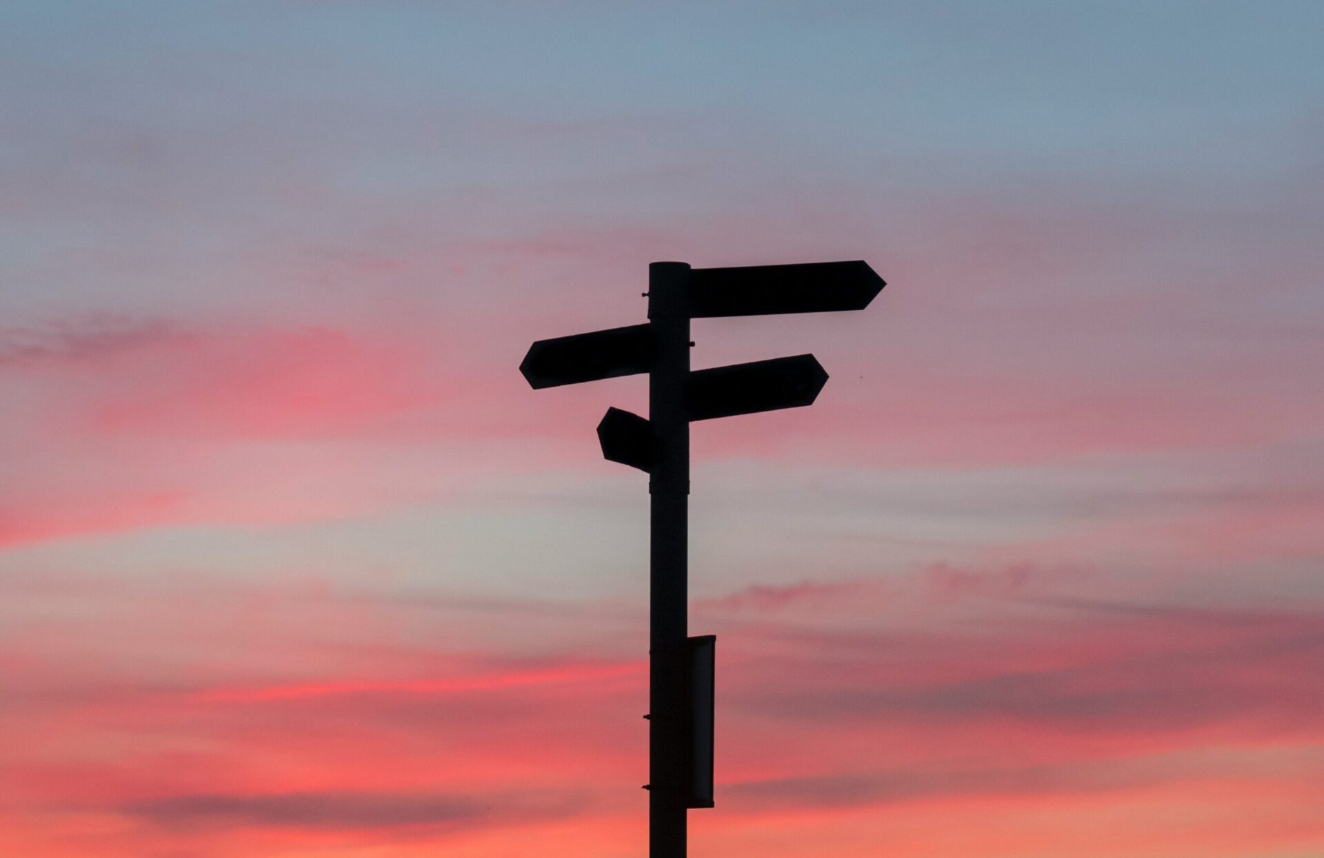 The image shows a silhouette of a signpost with multiple directional arrows against a colorful sunset sky. The sky has shades of pink, orange, and blue.