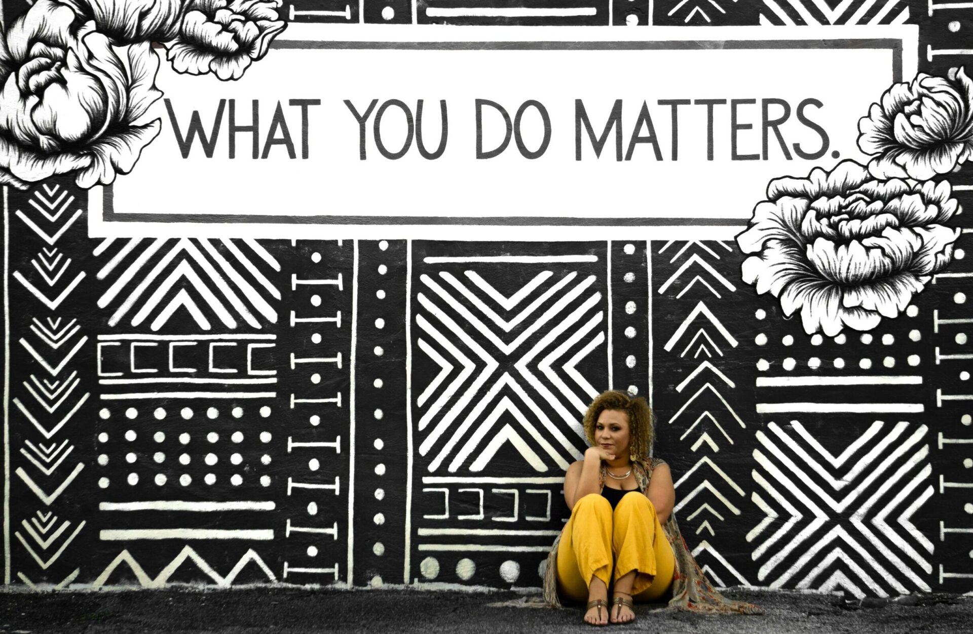 The image shows a mural with the text WHAT YOU DO MATTERS. The background has a black and white geometric and floral design. There is a person sitting in front of the mural, wearing yellow pants.